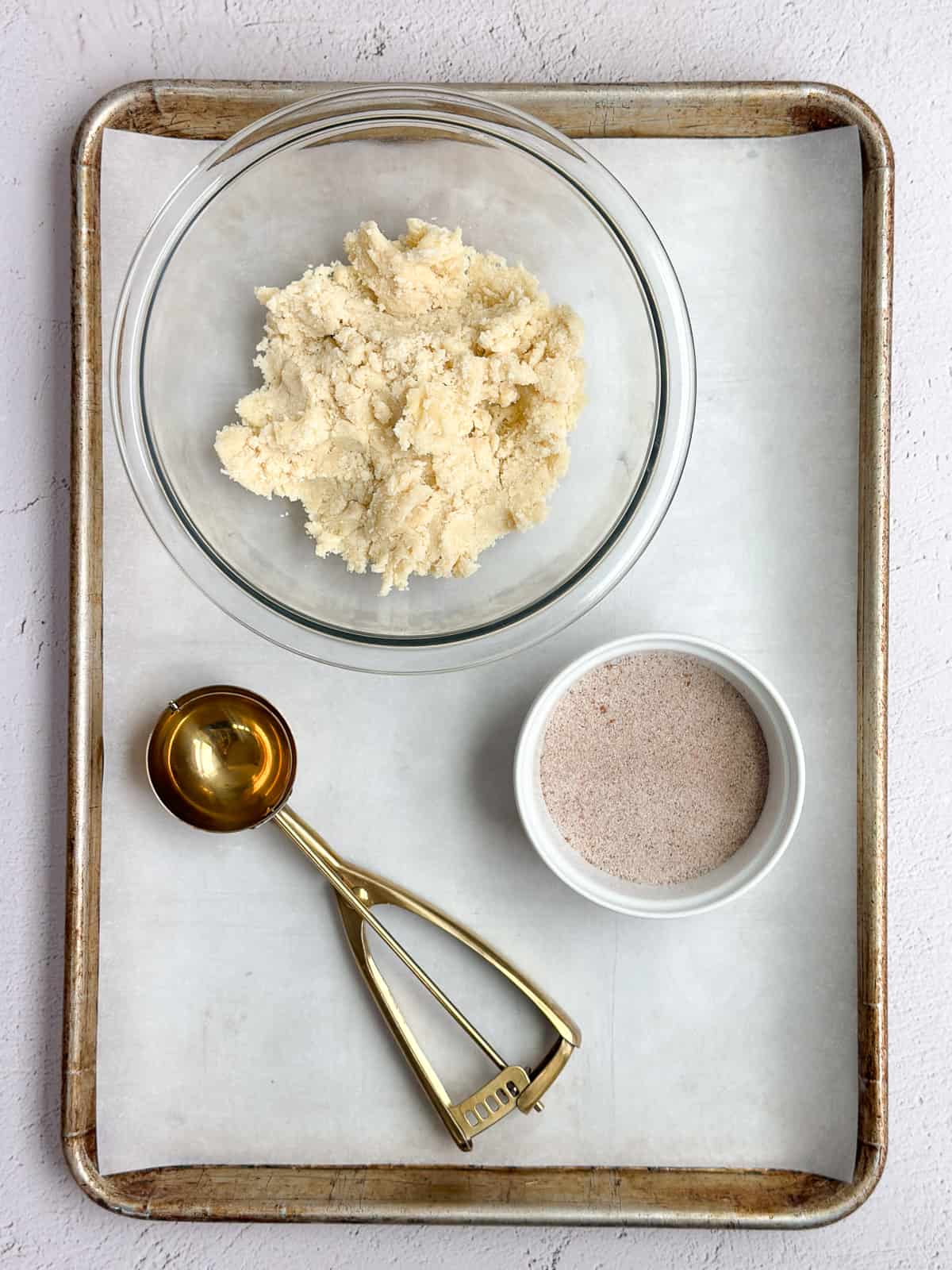 the cookie dough, cinnamon and sugar, cookie scoop and cookie sheet all stacked together.
