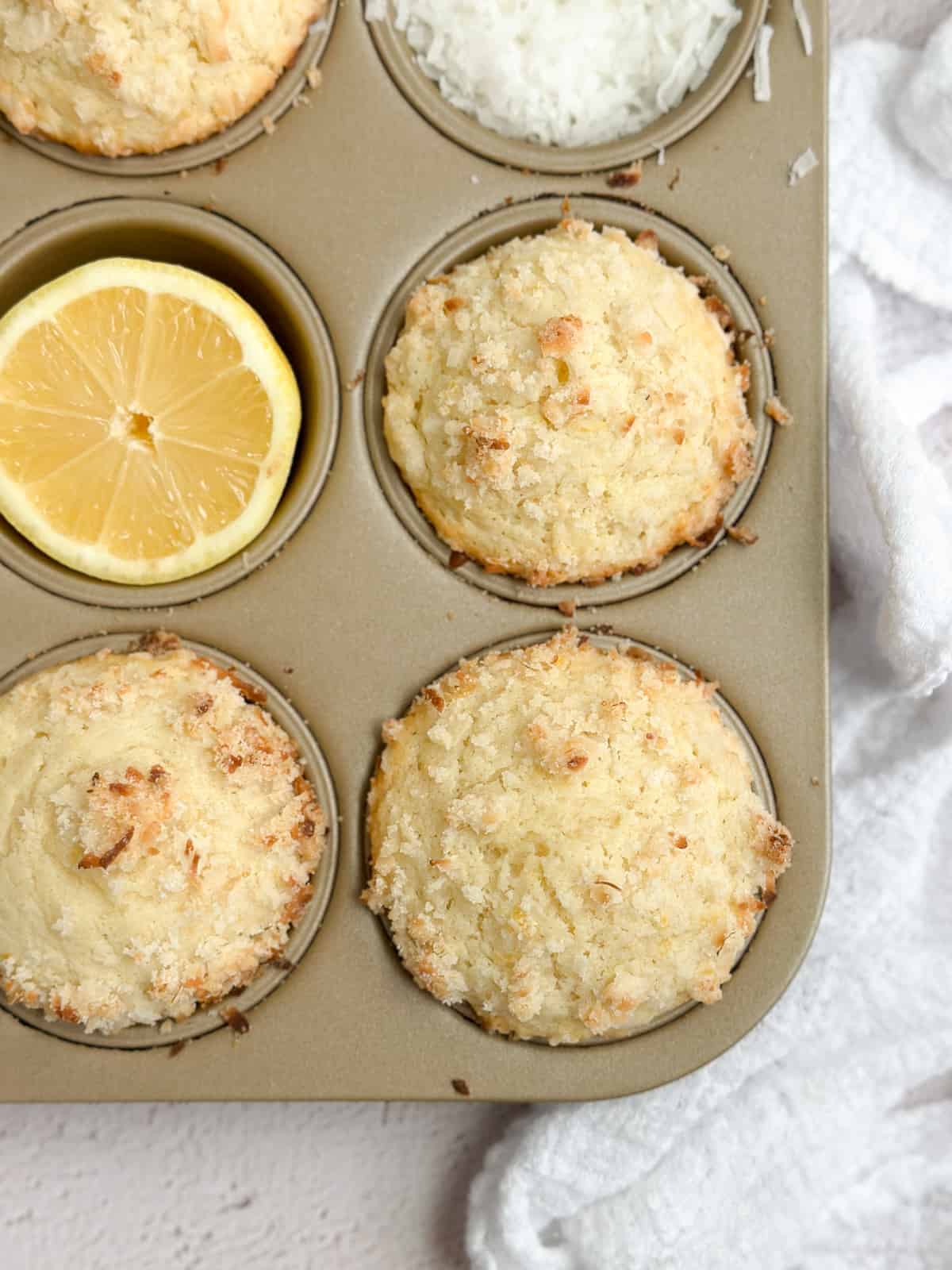 a few coconut lemon muffins in the muffin tin with a cut lemon and some coconut flakes.