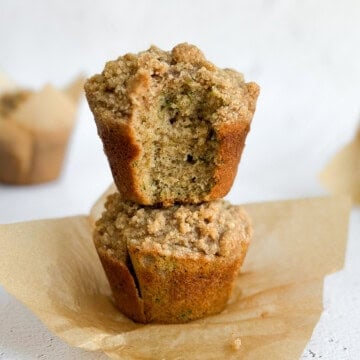 two cinnamon zucchini crumb muffins stacked on top of each other with a bite missing from the top one.