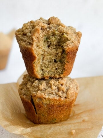 two cinnamon zucchini crumb muffins stacked on top of each other with a bite missing from the top one.