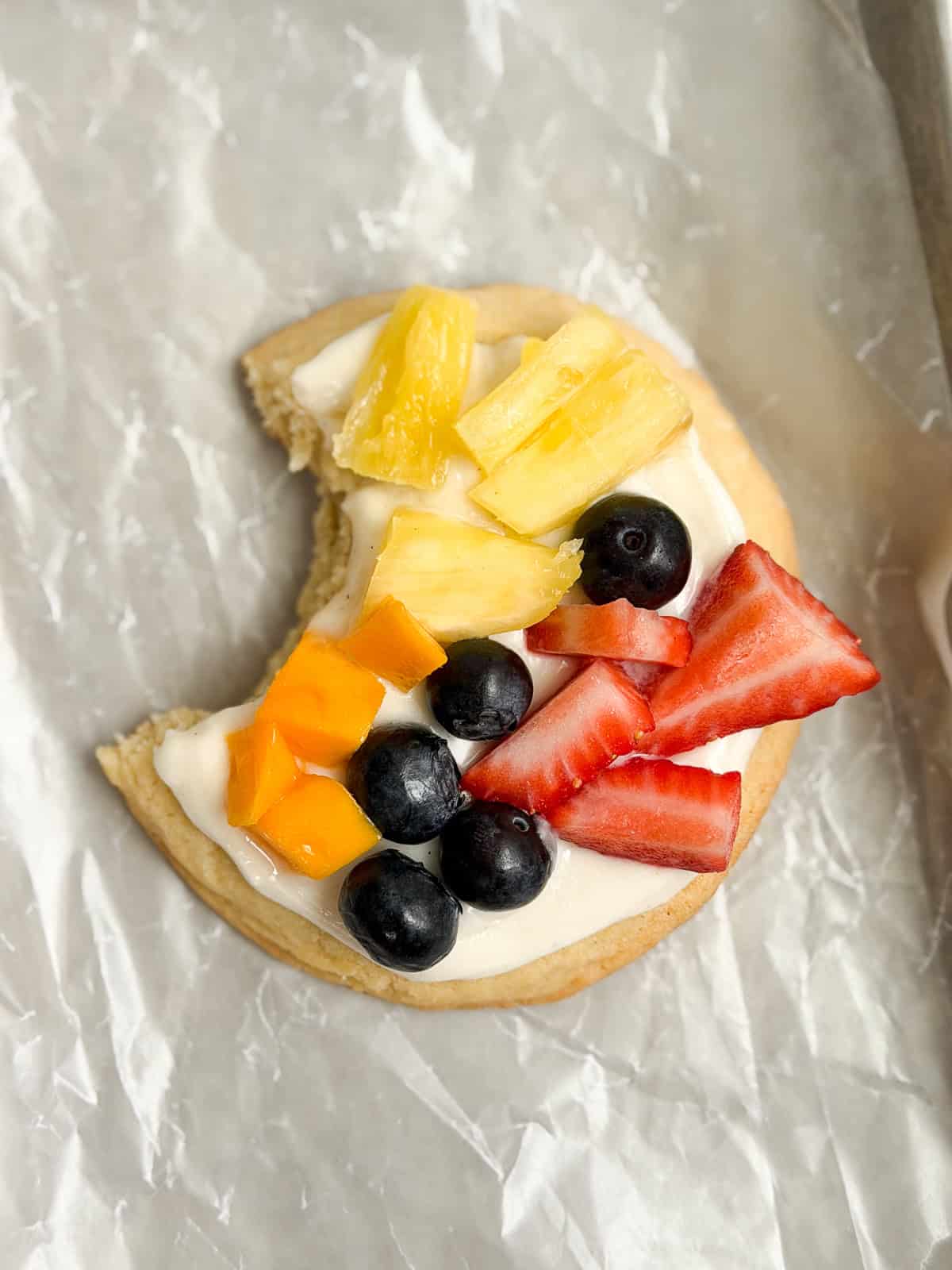 a single sugar cookie fruit pizza cookie with a bite missing.