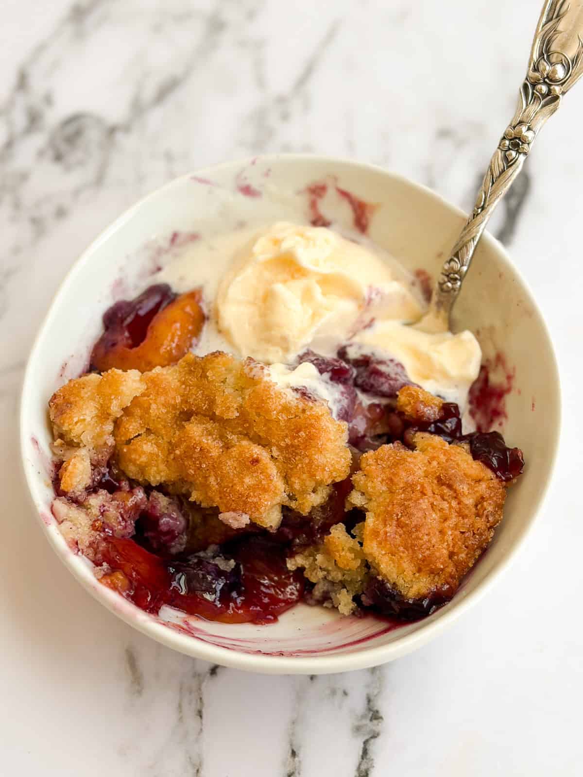 a bowl of blueberry peach cobbler with a scoop of ice cream and a spoon.